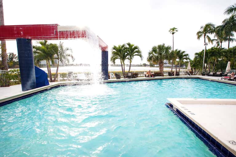 Resort pool with water slide feature, palm trees, and waterfront views on sunny day