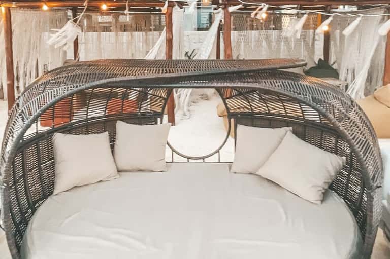 Curved woven daybed with white cushions and pillows on covered patio with hanging curtains