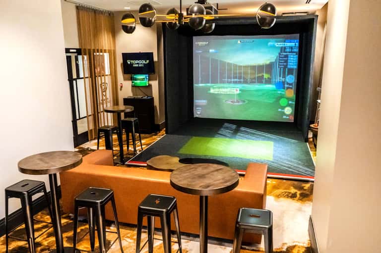 Indoor golf simulator bay with large screen, hitting mat, seating area, and game display