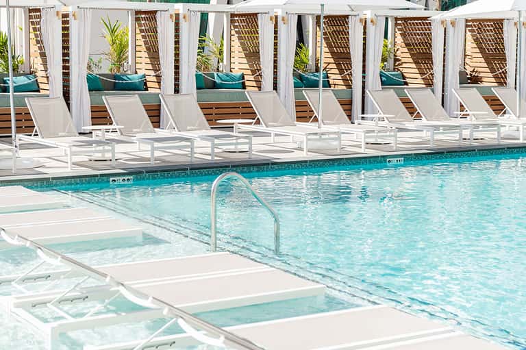 Resort pool with white lounge chairs and private cabanas with teal cushions and drapes