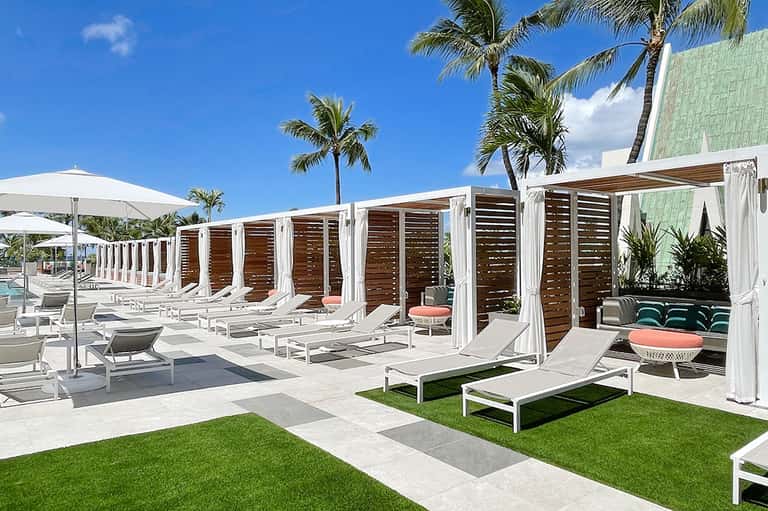 Modern resort pool deck with white cabanas, loungers, and palm trees under blue sky
