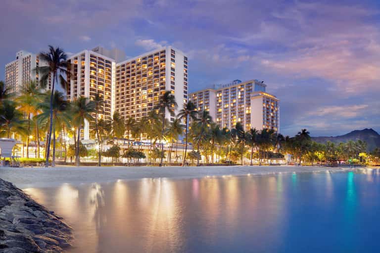 Beachfront resort buildings illuminated at dusk with palm trees and calm ocean waters