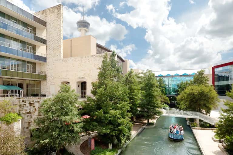Outdoor urban waterway with boat passing under bridge, surrounded by modern buildings, green trees, and stone architecture.