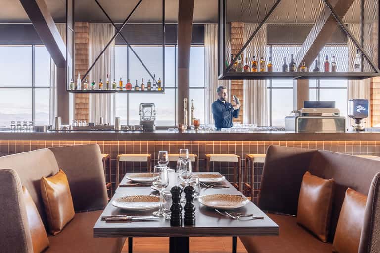 Modern restaurant bar with leather seating, hanging bottle display, and ocean views