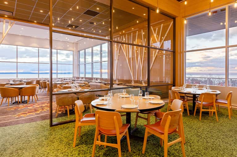 Upscale dining room with panoramic waterfront views, round tables with orange chairs, and floor-to-ceiling windows