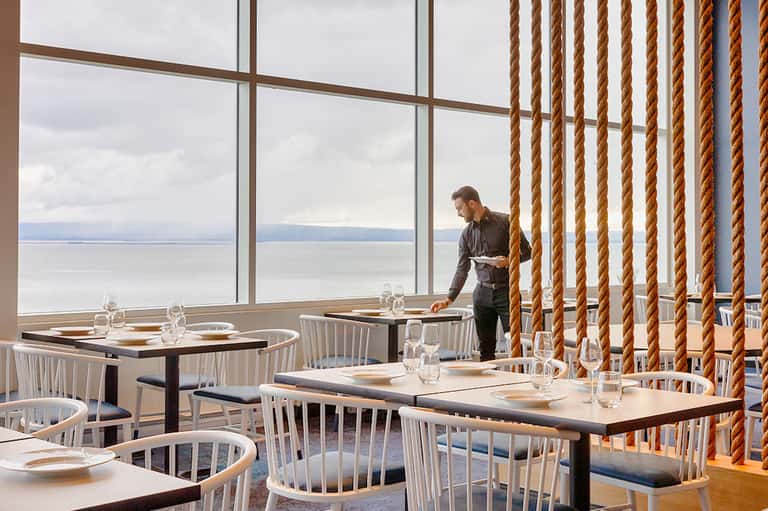 Modern restaurant dining area with floor-to-ceiling windows overlooking calm ocean waters and rope curtains