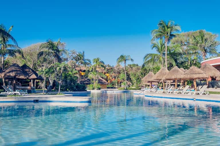 Resort pool with thatched-roof cabanas, palm trees, and ocean views