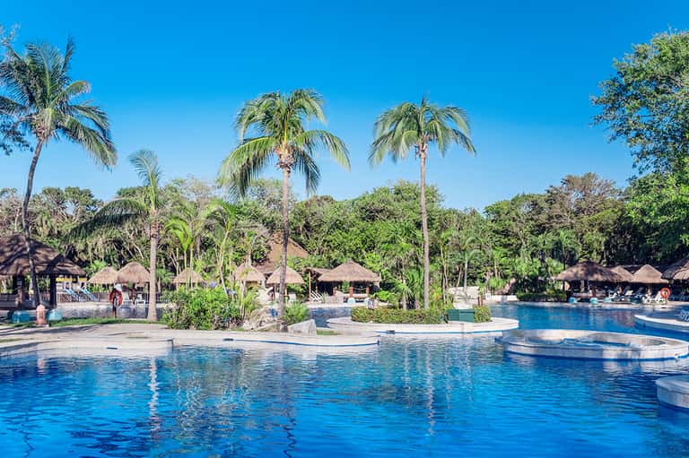 Tropical resort pool with palm trees, thatched-roof cabanas, and clear blue water surrounded by lush landscaping