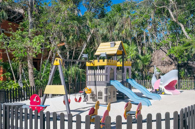 Children's playground with yellow and gray castle structure, blue slides, and swings in shaded area