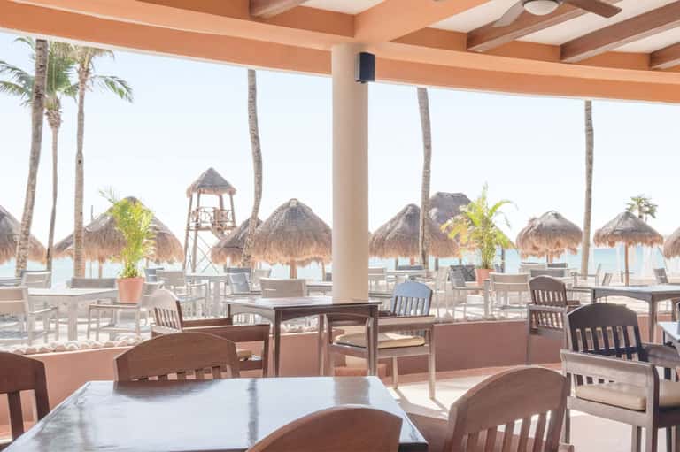 Beachfront restaurant dining area with ocean view, palm trees, thatched umbrellas, and casual seating