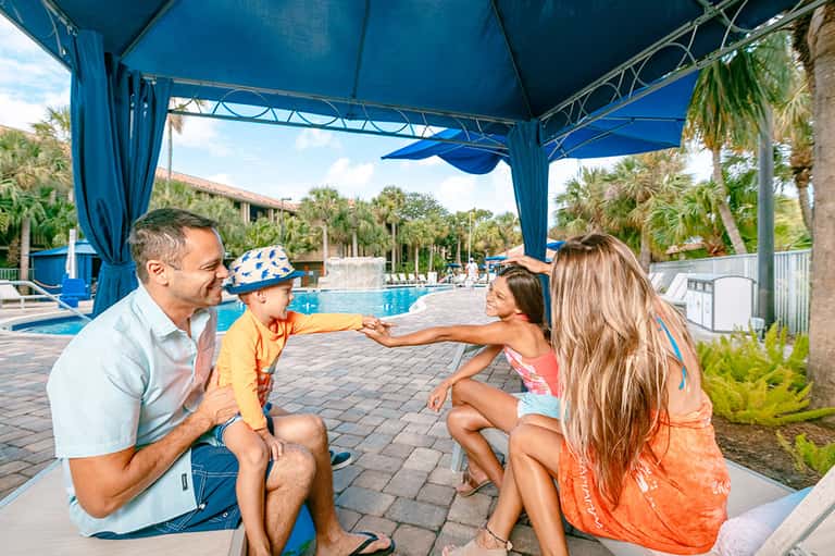 Family with young child under blue canopy by resort pool with palm trees and loungers