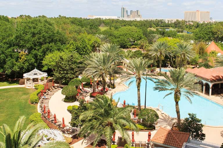 Aerial view of resort pool complex with palm trees, loungers, and downtown city skyline in background