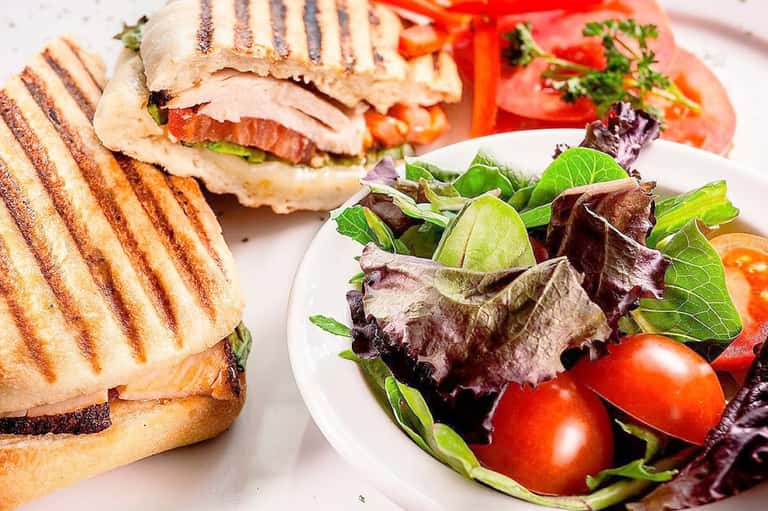 Grilled sandwich with fresh salad on white plate featuring tomatoes, greens, and herbs