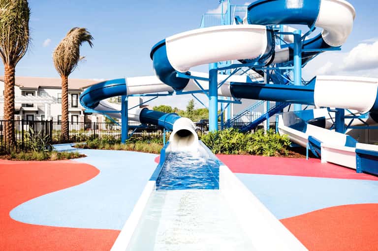 Water park with blue and white slide structures on red and blue padded surface near white residential buildings