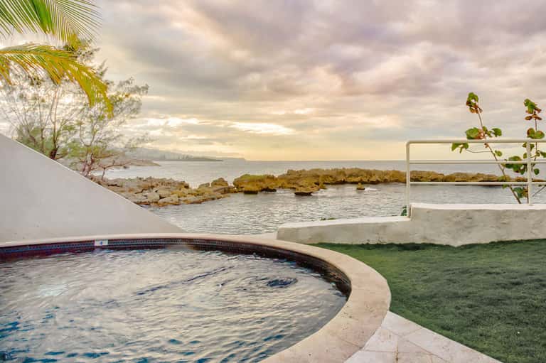 Curved hot tub overlooking ocean and rocky coastline with palm trees and cloudy sky