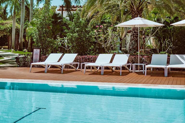 Resort pool deck with white lounge chairs, umbrellas, tropical palms, and lush landscaping