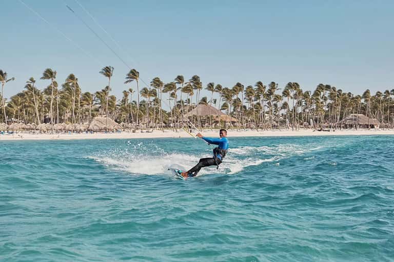 Kitesurfer in blue wetsuit riding waves with palm tree-lined beach and resort buildings in background