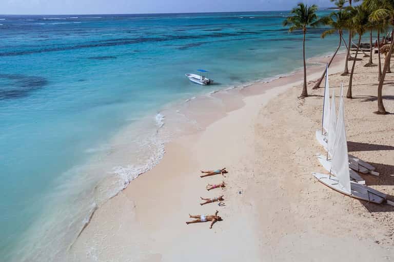 Pristine turquoise beach with palm trees, sailboats, and guests relaxing on sand