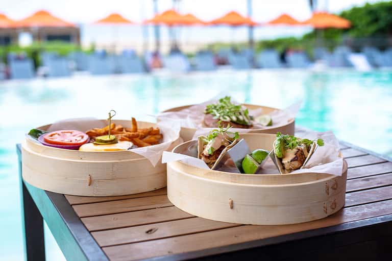 Bamboo steamer baskets with gourmet food and fries on a wooden deck overlooking a resort pool