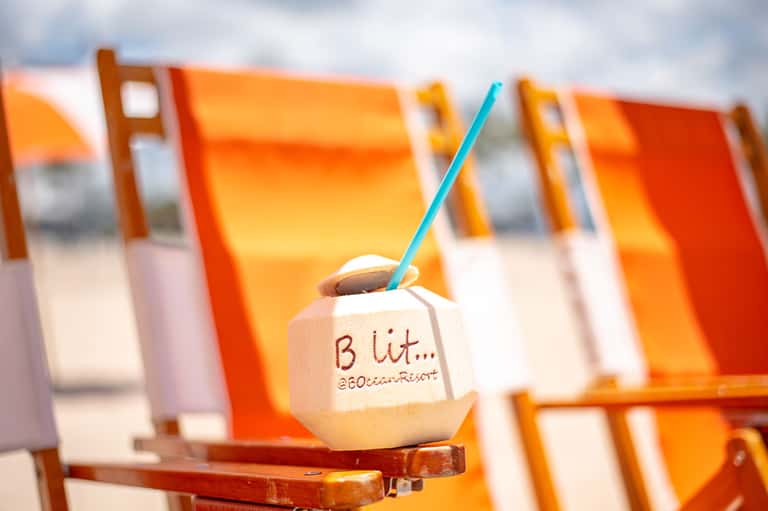 Coconut drink with blue straw on beach chair with orange loungers blurred in background