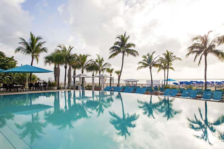 Resort infinity pool with palm trees and lounge chairs overlooking distant mountains at dusk