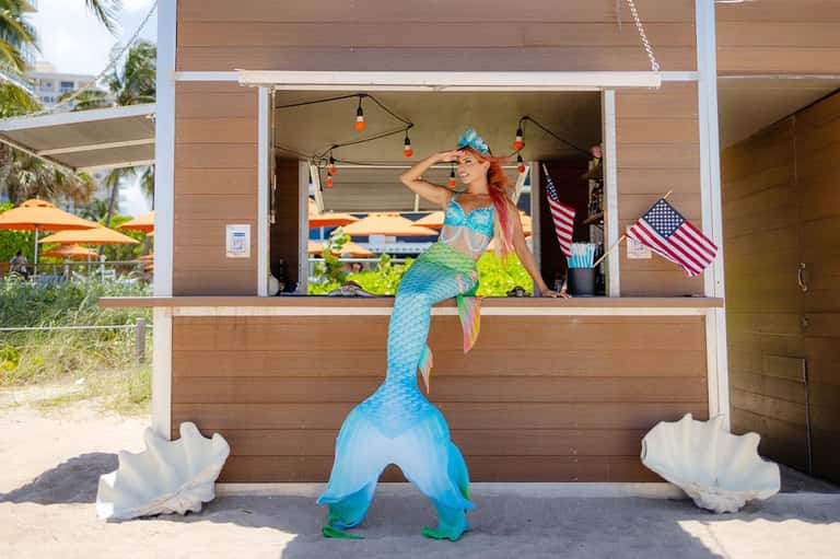 Mermaid performer at beachside tiki bar with surfboards and American flags