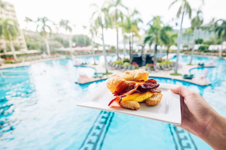 Breakfast sandwich with bacon and egg held over resort swimming pool with palm trees