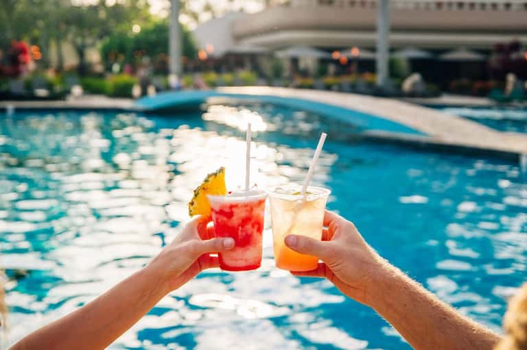 Two colorful cocktails with straws held up by a pool with tropical lounge chairs and umbrellas in background