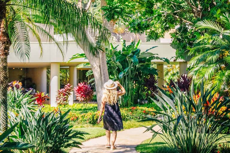 Woman in black dress and white hat walking through lush tropical garden courtyard