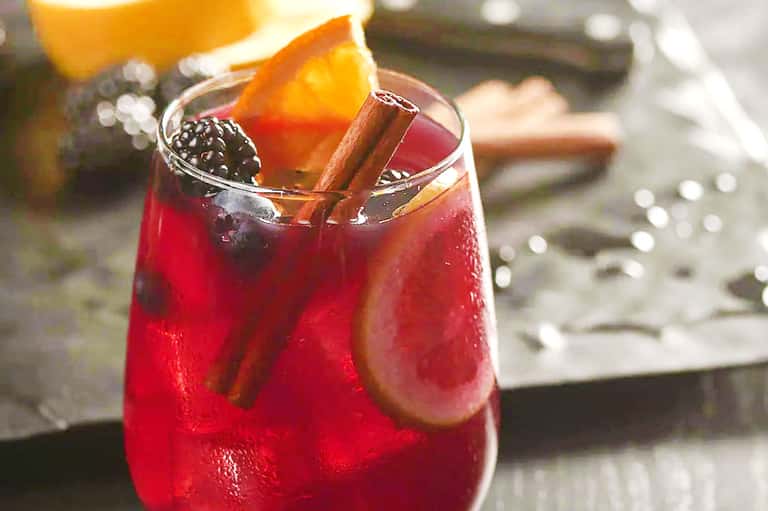 Specialty cocktail with cinnamon stick, fresh orange slice, and blackberry garnish on white surface