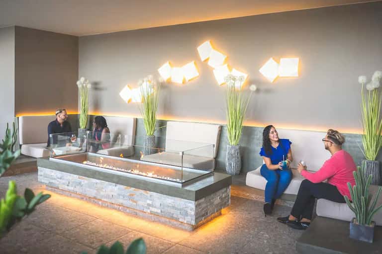 Modern hotel lobby with fireplace, decorative lighting, tall floral arrangements, and guests in seating area