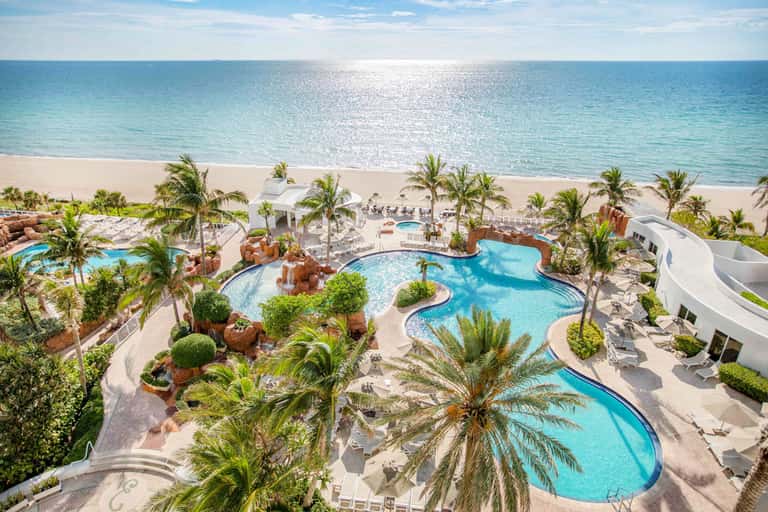 Aerial view of beachfront resort with multiple curved pools, palm trees, white buildings, and pristine sandy beach