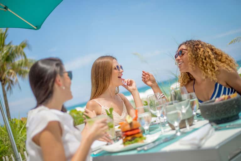 Three women dining at a beachfront restaurant under a green umbrella with ocean view