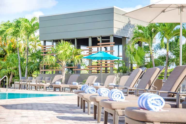 Luxury resort pool area with lounge chairs, blue umbrellas, palm trees, and modern architecture building