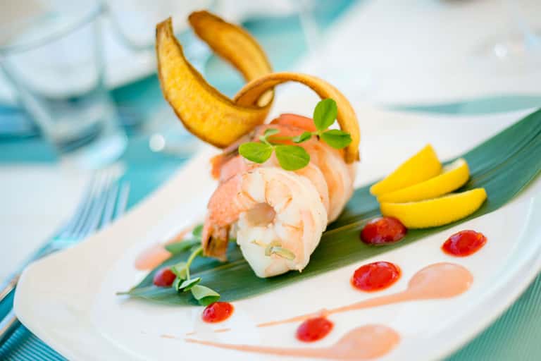 Elegantly plated shrimp dish with banana garnish and red sauce on green leaf, fine dining presentation