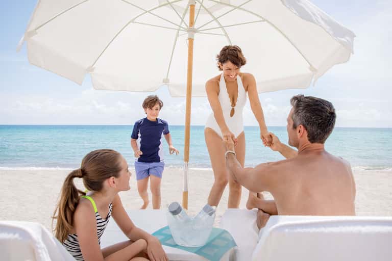 Family relaxing under white beach umbrella with turquoise ocean and sandy beach in background