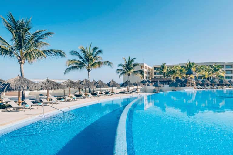 Large resort pool with palm trees, umbrellas, loungers, and beachfront hotel building