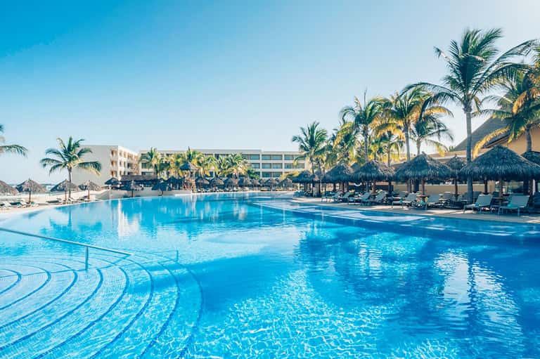 Large resort pool with turquoise water, tiki umbrellas, lounge chairs, and palm-lined beachfront