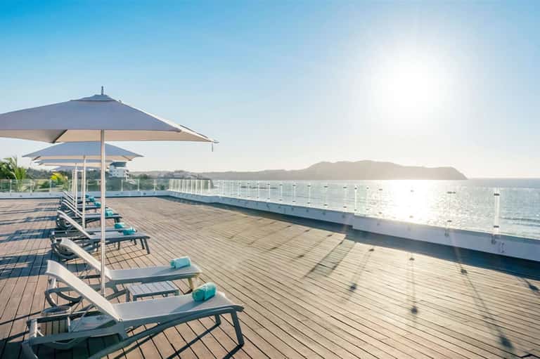 Oceanfront deck with white loungers and umbrellas overlooking a calm bay with distant mountains