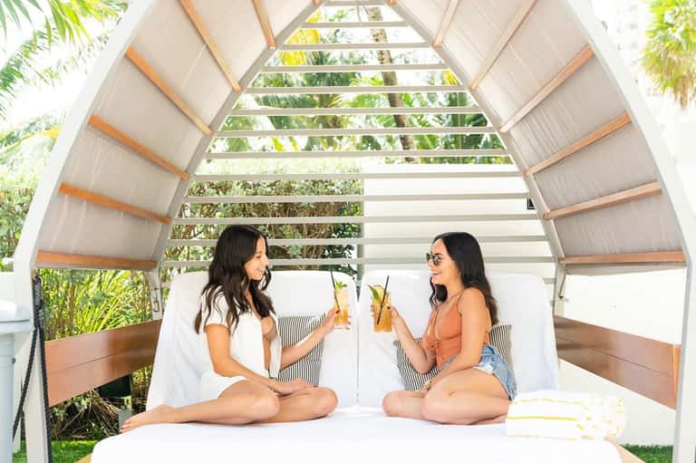 Two women in a cabana with arched roof sharing drinks among tropical plants