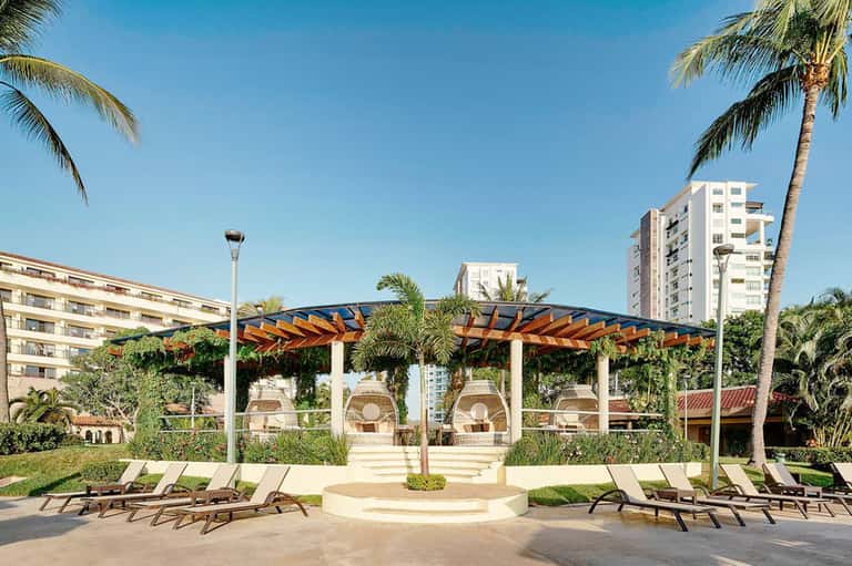 Resort courtyard with wooden pergola, lounge chairs, palm trees, and multi-story hotel buildings