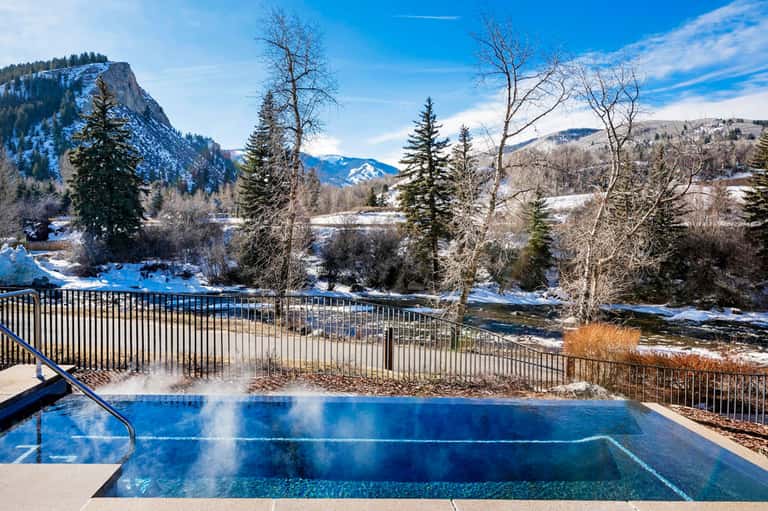 Steaming hot spring pool with mountain and river views, surrounded by snow and evergreen trees