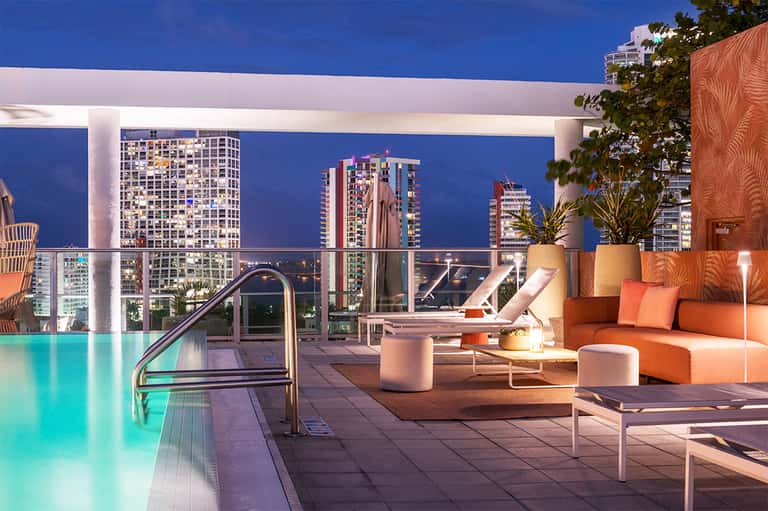 Illuminated rooftop pool and lounge area at night with city skyline views and orange seating