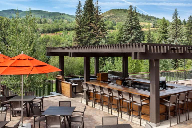 Outdoor mountain resort terrace with dark pergola, wooden bar, orange umbrella, and forested hillside views
