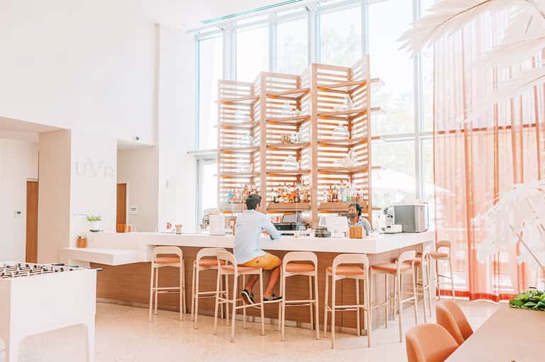Contemporary hotel bar with wooden shelving, white seating, and large windows