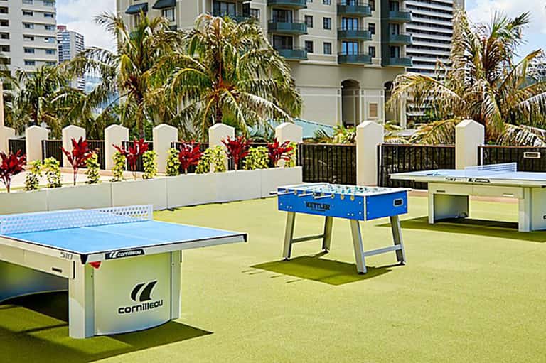 Rooftop recreation area with ping pong and foosball tables, white fence, palm trees, and city skyline