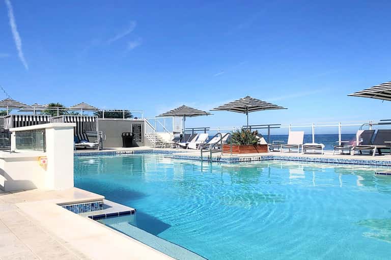 Resort pool with clear turquoise water, white umbrellas, loungers, and ocean view under blue sky