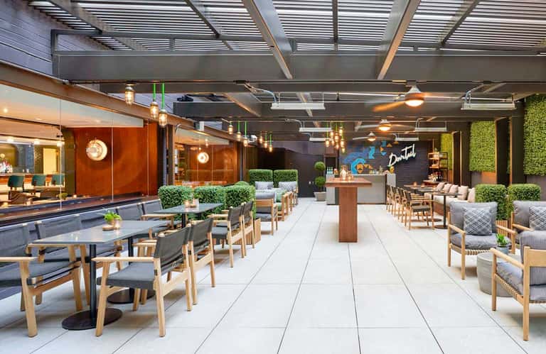Contemporary hotel terrace with wooden tables, chairs, hanging lights, and glass storefront