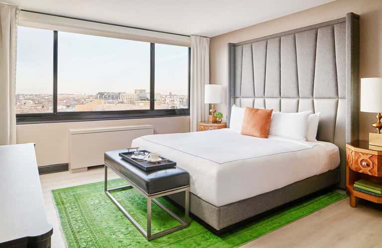 Modern hotel bedroom with city skyline view, neutral bedding, orange accent pillow, and green runner rug