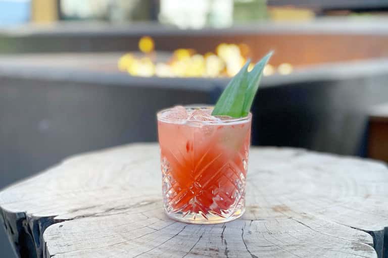 Coral-colored cocktail with ice and green leaf garnish served on wooden surface